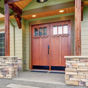 Wooden double entry doors with windows on the top third on the exterior of a greenish house and wood accents.